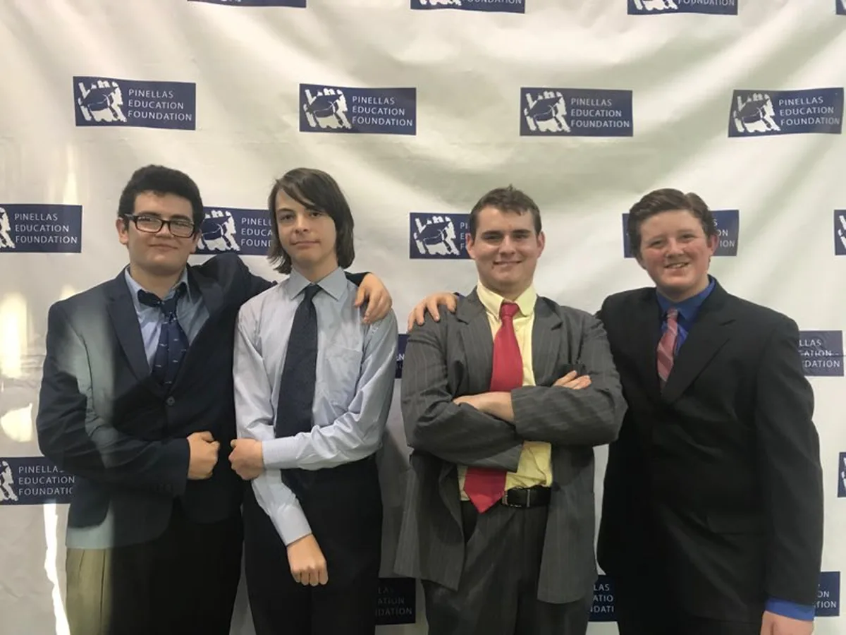 Four Next Gen Tech students in suits, arms around each other, in front of a step-and-repeat banner printed with the Pinellas Education Foundation logo.