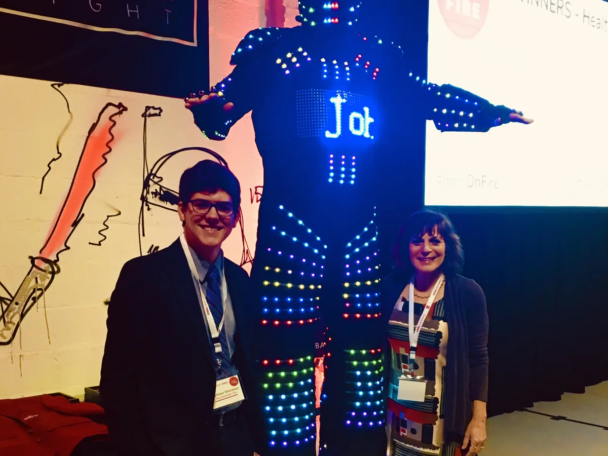A program student and Robyn Mussler standing either side of a tall LED-covered stilt-walker costume at an innovation awards event.